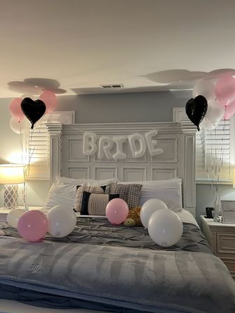Cozy master bedroom bridal surprise with white 'BRIDE' letter balloons and pink, white, and black heart balloons floating around a neatly made gray bed.