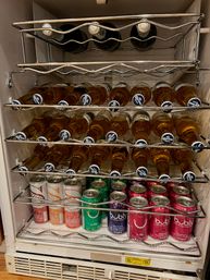 Compact under-counter beverage fridge stocked with rows of chilled Corona beer bottles on chrome racks and colorful Bubly sparkling water cans on the bottom shelf