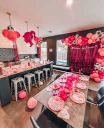 Barbie-themed bachelorette party setup in a modern home kitchen and dining area with pink and cow-print balloon garlands, metallic pink fringe backdrop reading “Barbie’s Last Rodeo,” barstool counter, themed place settings with pink feather boas and scattered balloons.
