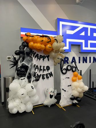 Spooky Halloween balloon display with orange, black and white balloons, cobwebs, bat cutouts, dripping 'Happy Halloween' letters and a smiling ghost foil set up inside an indoor gym training area.