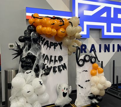 Spooky Halloween balloon display with orange, black and white balloons, cobwebs, bat cutouts, dripping 'Happy Halloween' letters and a smiling ghost foil set up inside an indoor gym training area.
