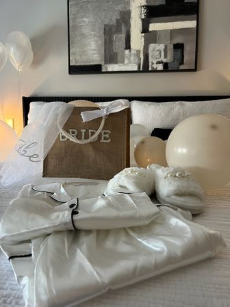 Wedding morning bed setup with a burlap bag reading 'BRIDE' draped with a veil and ribbon, white satin robe with black trim, pearl‑embellished fuzzy slippers and cream balloons beneath abstract wall art.
