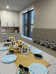 Modern home kitchen with a kids' party table: gold glitter runner, light blue plates, framed black-and-white child photos with paper crowns, sunflower centerpieces, sunglasses favors and repeated smiling face cutouts along the wall.