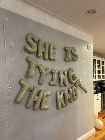 Gold foil letter balloons reading "SHE IS TYING THE KNOT" strung on a gray interior wall above hardwood floor near white kitchen cabinets — indoor bridal shower/home celebration decor.