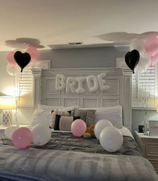 Cozy master bedroom decorated for a bridal celebration with white inflatable 'BRIDE' letters over a white headboard, pink, white and black heart balloons tied at each side and scattered on a gray bed with decorative pillows.