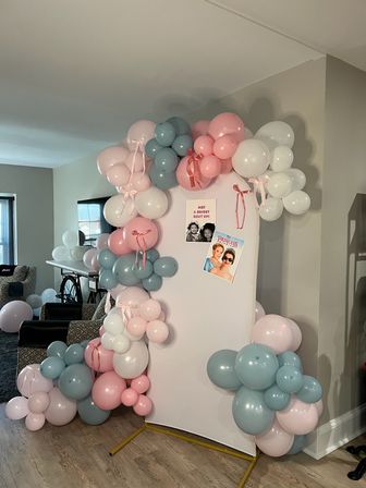 Playful pastel balloon arch in pink, dusty blue and white with ribbon bows framing a freestanding photo backdrop in a living room—stylish indoor party decor for a birthday or baby shower.