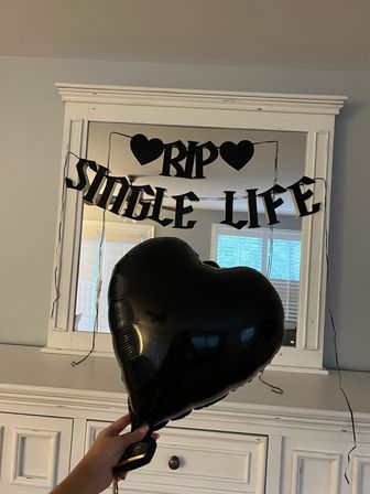 Black heart-shaped balloon held in front of a white dresser and mirror decorated with a black "RIP Single Life" banner — indoor bachelorette/bridal party decor.