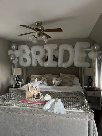 Bridal bedroom setup with oversized white letter balloons spelling "BRIDE" above a bed, silver heart and helium balloons, gift bag, veil and sash arranged on a tufted bedspread.