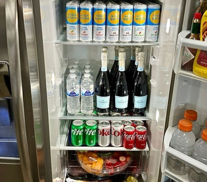 Open stainless-steel kitchen refrigerator neatly stocked with canned hard seltzers on the top shelf, bottled water and sparkling wine on the middle shelf, soda cans and pre-cut fruit bowls below, and sports drinks in the door.