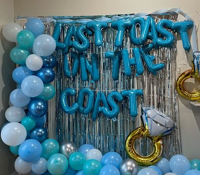 Blue and teal balloon garland framing a silver fringe backdrop with blue foil letter balloons forming a celebration phrase and a gold diamond-ring foil balloon — festive photo‑booth setup.