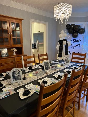 Black-and-white themed home dining room party setup with a long table dressed with framed photos, candles, napkins, gift bags, a balloon arch and crystal chandelier, kitchen visible in the background.