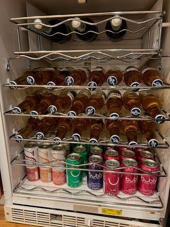 Compact under-counter beverage fridge stocked with rows of chilled Corona beer bottles on chrome racks and colorful Bubly sparkling water cans on the bottom shelf