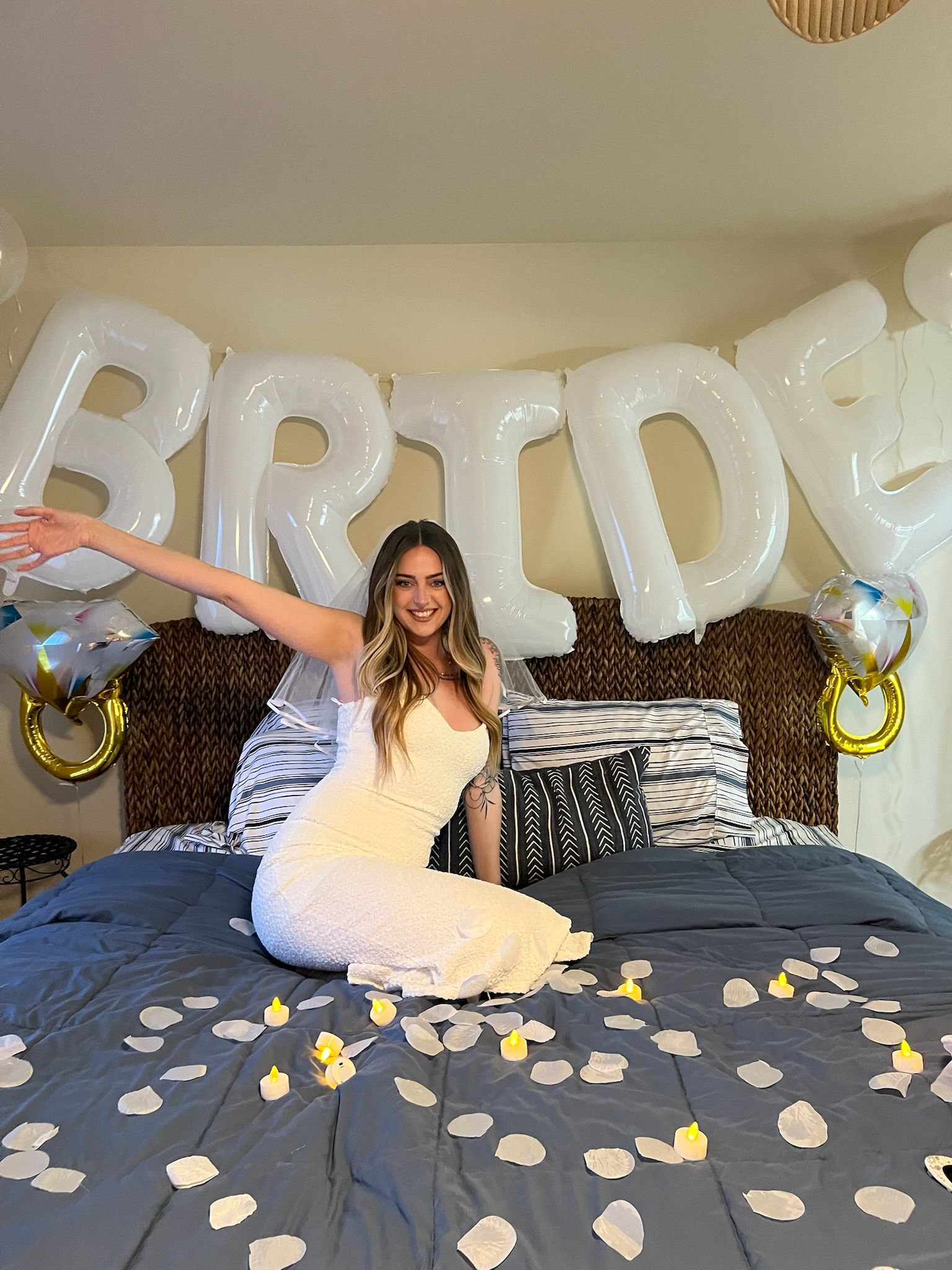 Bride-to-be in a white dress sitting on a bed under large white 'BRIDE' balloons with faux rose petals and LED candles for an indoor bridal shower celebration.