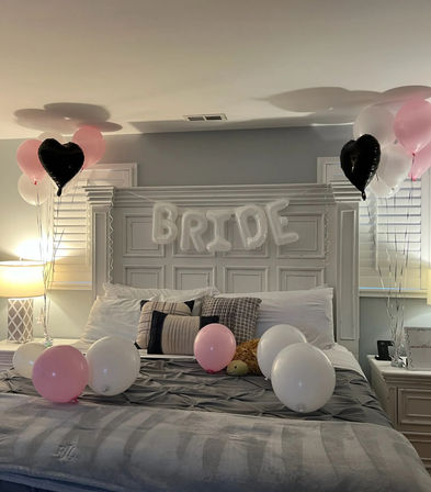 Cozy master bedroom decorated for a bridal celebration with white inflatable 'BRIDE' letters over a white headboard, pink, white and black heart balloons tied at each side and scattered on a gray bed with decorative pillows.