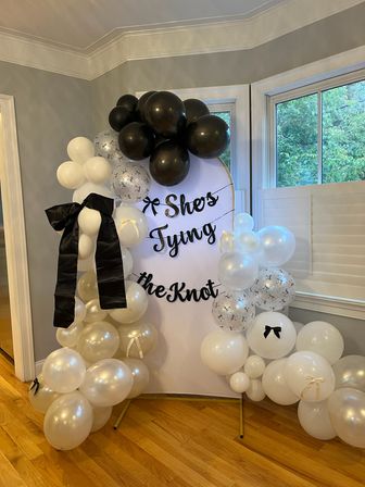 Indoor bridal shower backdrop in a home with a white sign reading She's Tying the Knot, surrounded by black, white and pearl balloons, confetti balloons and a large black bow on a hardwood floor near windows.