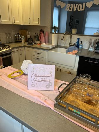 Pink-themed brunch setup on a modern home kitchen island with a “Champagne Problems” sign, champagne coupe glasses on a wooden tray, pink cups and paper decorations, farmhouse sink and a LOVE banner in the background