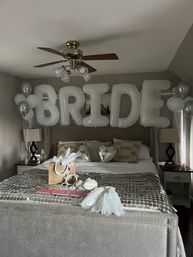 Neutral-toned bedroom styled as a bridal suite with large white letter balloons spelling "BRIDE" above the bed, silver heart and cluster balloons, and bridal accessories (veil, sash, gift bag) arranged on the bed.
