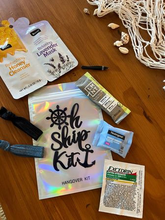 Nautical hangover kit on wooden table with seashells: holographic "Oh Ship Kit" pouch, honey cleanser and lavender face masks, electrolyte stick, makeup wipe, pain-relief sachet and hair ties.