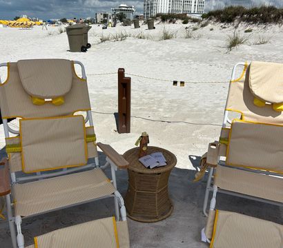 Pair of yellow-striped beach chairs and wicker side table with sunscreen on a white-sand beach, stormy gray clouds overhead and beachfront condos, trash bins and yellow umbrellas in the distance.