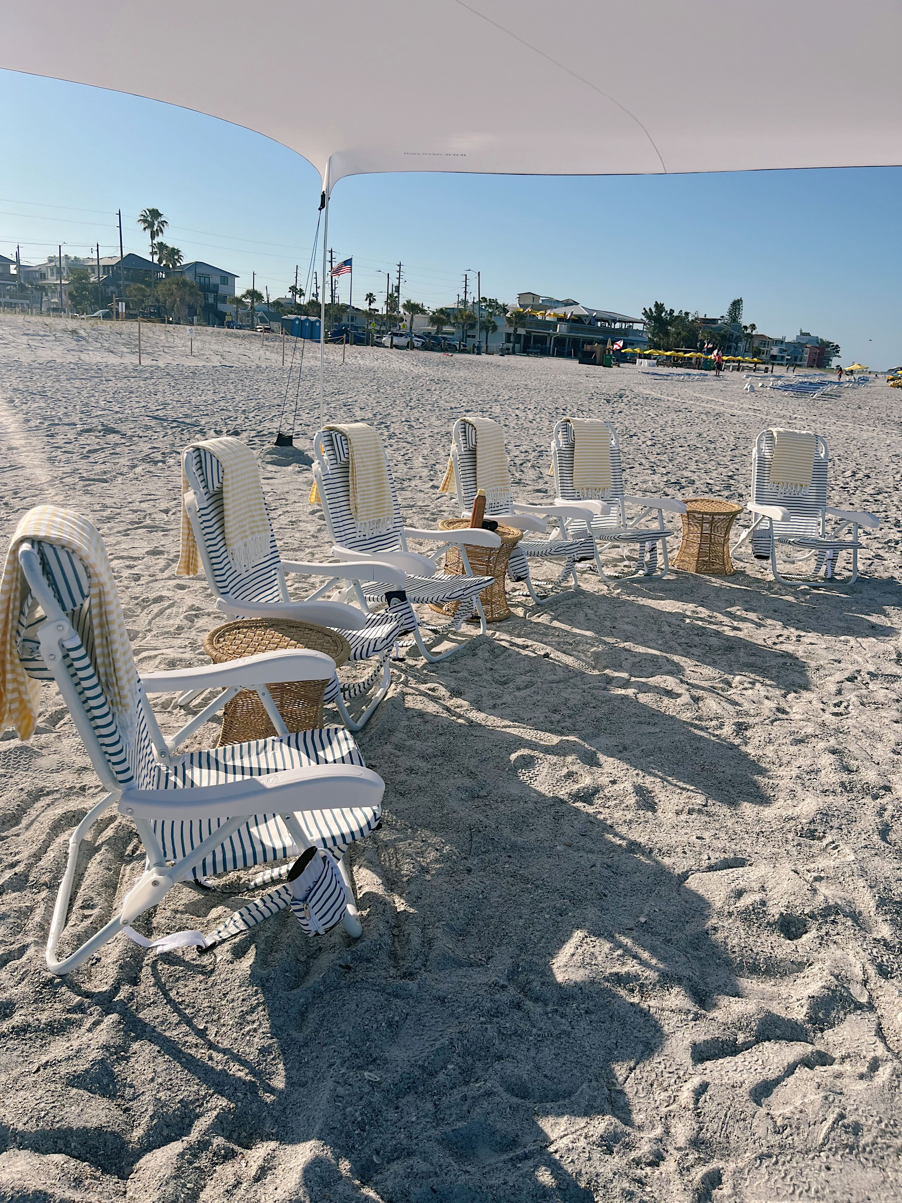 Striped lounge chairs lined up under a white canopy on a sunny sandy beach with yellow towels and wicker side tables, palm trees and beachside buildings along the coast under a clear blue sky.