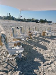 Striped lounge chairs lined up under a white canopy on a sunny sandy beach with yellow towels and wicker side tables, palm trees and beachside buildings along the coast under a clear blue sky.