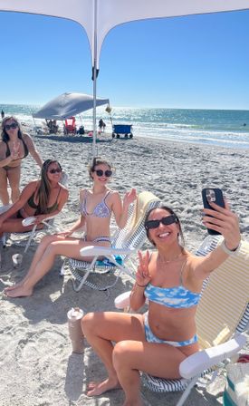 Smiling friends in bikinis taking a selfie under a beach canopy on a sunny sandy oceanfront with sparkling waves