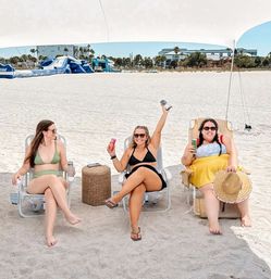 Three friends in swimsuits relax under a beach canopy on white sand, sitting in folding chairs with canned drinks and a wicker side table, with an inflatable water park, palm trees and resort buildings along the shoreline in the background.