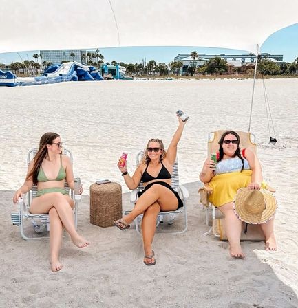 Three friends in swimsuits relax under a beach canopy on white sand, sitting in folding chairs with canned drinks and a wicker side table, with an inflatable water park, palm trees and resort buildings along the shoreline in the background.