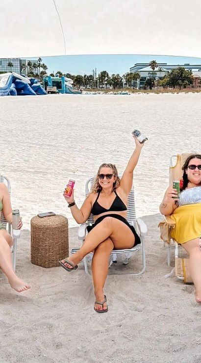 Three friends in swimsuits relax under a beach canopy on white sand, sitting in folding chairs with canned drinks and a wicker side table, with an inflatable water park, palm trees and resort buildings along the shoreline in the background.