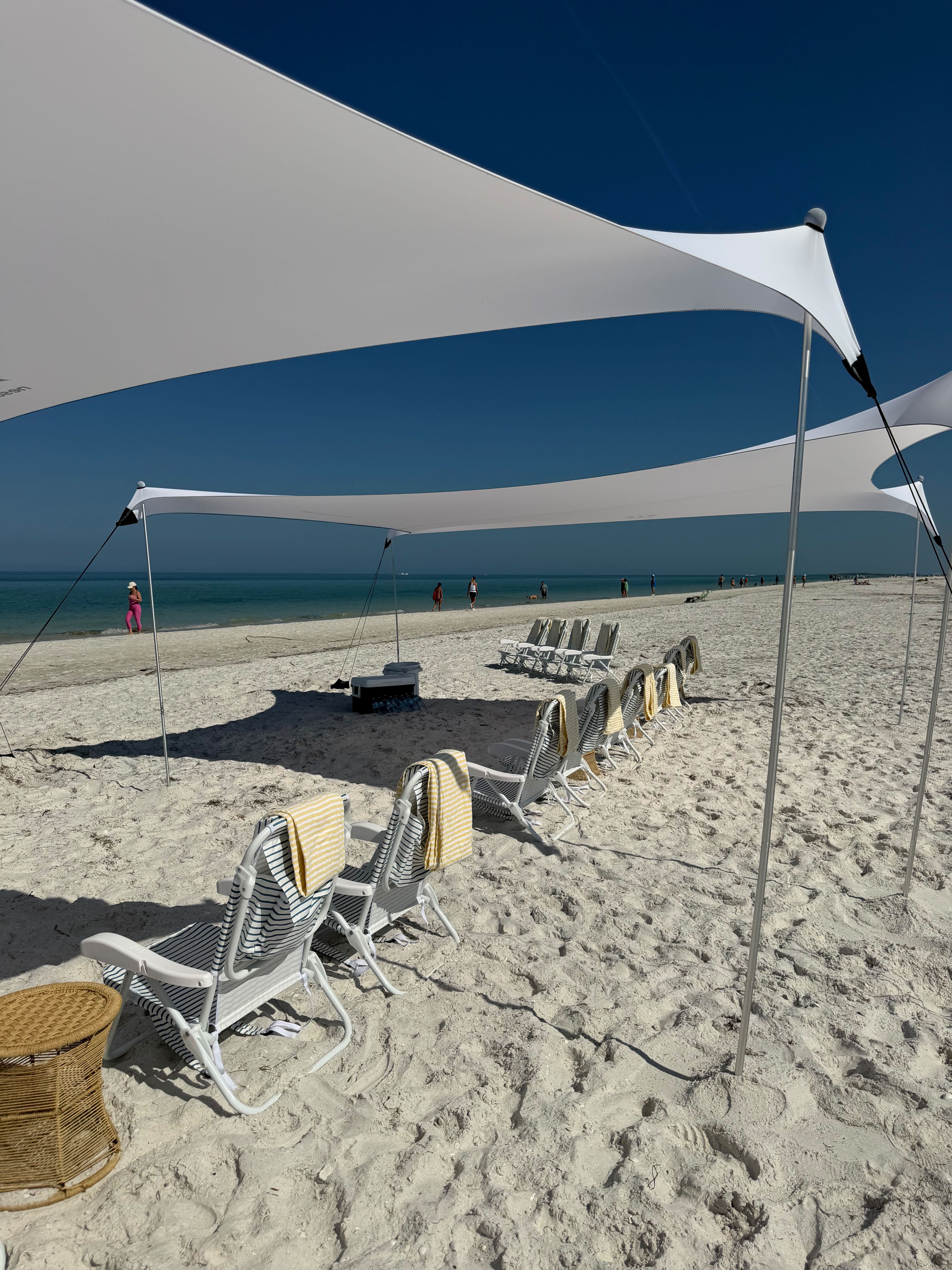White shade sails stretching over a row of striped lounge chairs with yellow towels on a sunny white-sand beach, turquoise water and deep blue sky with people walking along the shoreline in the distance.