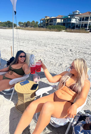 Two friends in bikinis clinking pink drinks under a beach umbrella on a sunny white‑sand beach with oceanfront homes and palm trees in the background.
