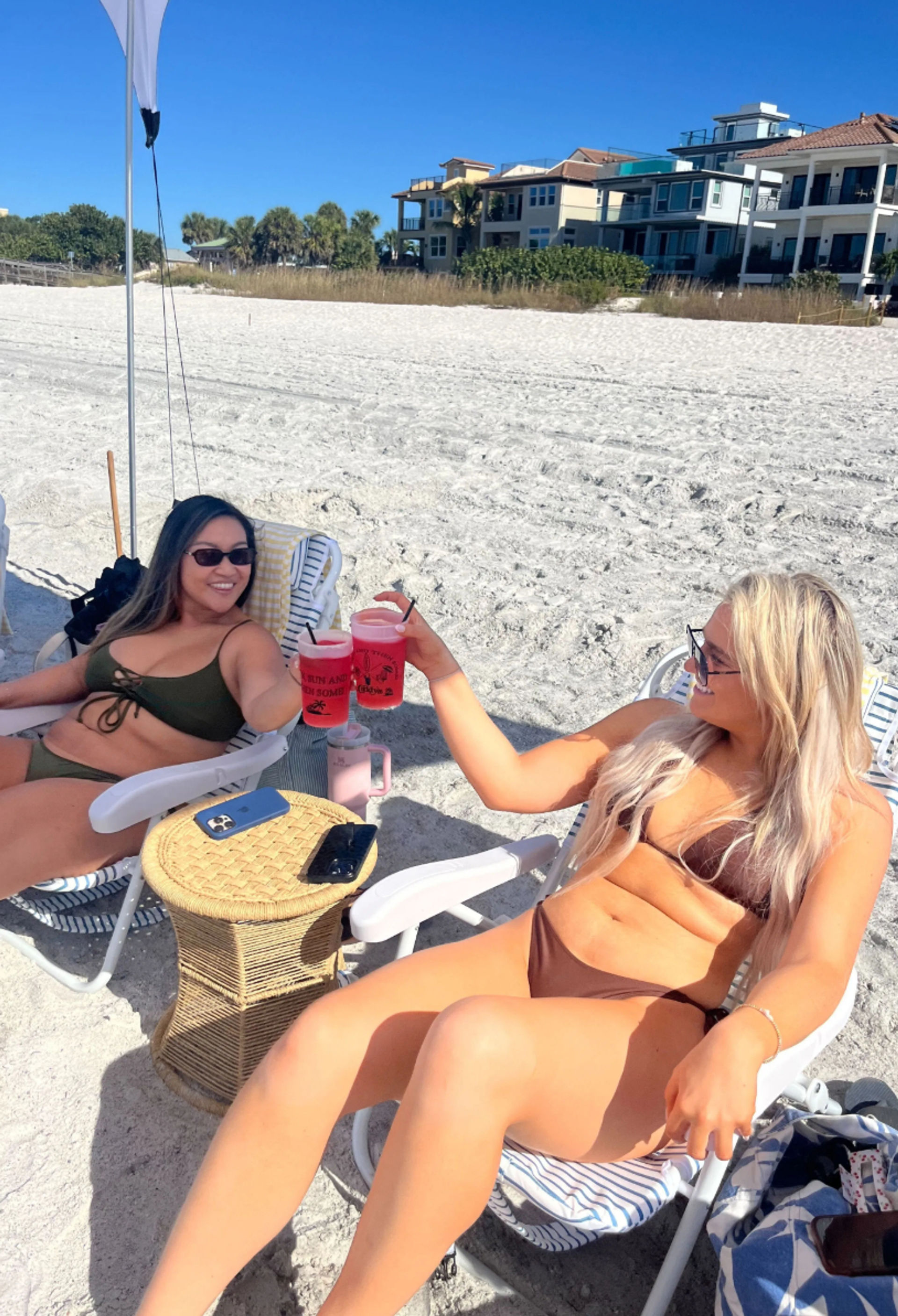 Two friends in bikinis clinking pink drinks under a beach umbrella on a sunny white‑sand beach with oceanfront homes and palm trees in the background.