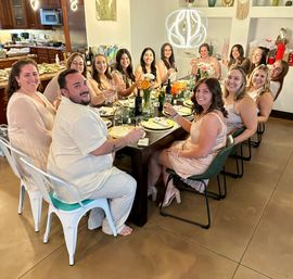 Group of friends toasting at a lively home dinner party around a long dining table with flowers, wine, and a modern pendant light, kitchen in the background.
