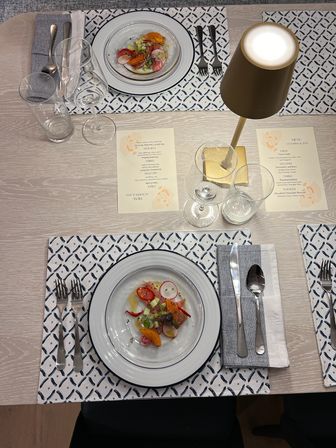 Top-down view of an indoor dining table set for two: white-rimmed plates with colorful citrus and radish salad, patterned placemats, folded gray napkins with cutlery, wine and water glasses, printed menu cards, and a small brass table lamp.