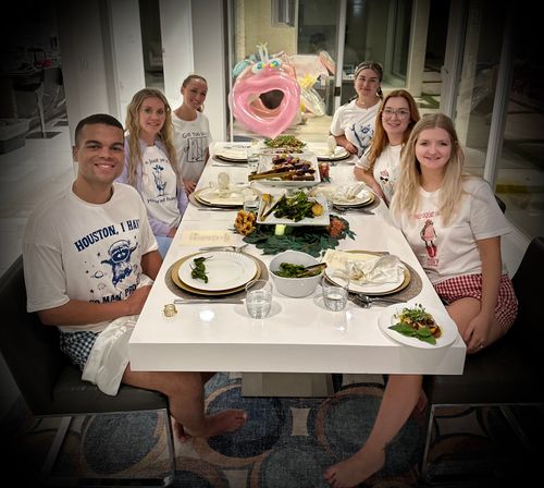 Seven friends in casual graphic tees gathered around a modern white dining table in a home dining room, enjoying plated appetizers and salads, with a large pink inflatable float visible in the background.