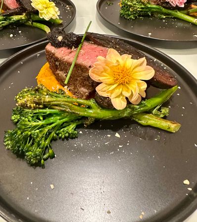 Medium-rare steak on a black plate with broccolini, orange puree, a dollop of dark sauce and a yellow edible flower garnish — elegant plated restaurant entrée.