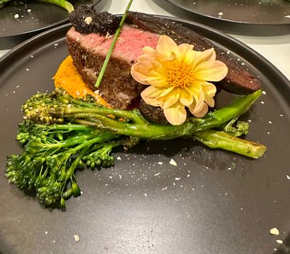 Medium-rare seared steak on orange vegetable purée with sautéed broccolini, truffle paste, chive and an edible yellow flower on a matte black plate — elegant fine-dining entree.