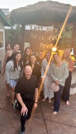 Smiling group of friends posing on a backyard patio under a thatched tiki hut at sunset, warm lamp glow and string lights framing a playful banner reading “LAST PICKLE SHE'LL EVER TICKLE” — casual outdoor party vibe.