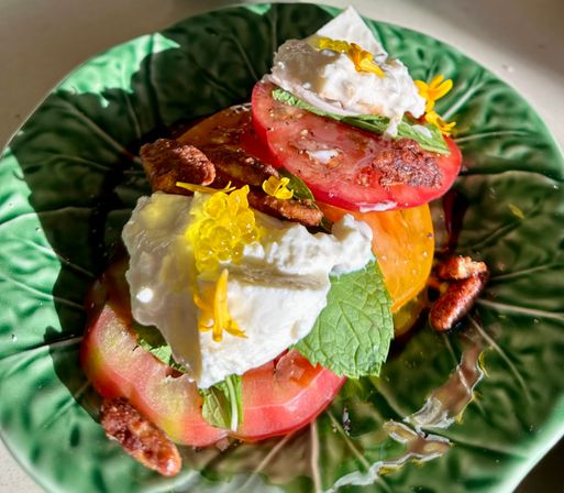 Sunlit heirloom tomato and burrata salad with basil and mint, candied pecans, edible yellow flowers and olive oil on a green leaf-pattern ceramic plate