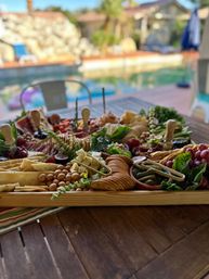 Sunlit poolside charcuterie board on a wooden tray with assorted cheeses, crackers, cured meats, grapes, cornichons, nuts, fresh herbs and breadsticks on a backyard patio table
