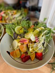 Fresh seasonal salad bowl with mixed greens, microgreens, sliced radishes and cucumbers, colorful tomato wedges and strawberries, citrus slices and vinaigrette with a balsamic drizzle in a white bowl on a wooden table — healthy summer lunch.