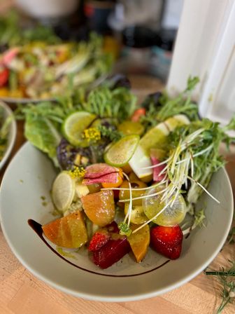 Fresh seasonal salad bowl with mixed greens, microgreens, sliced radishes and cucumbers, colorful tomato wedges and strawberries, citrus slices and vinaigrette with a balsamic drizzle in a white bowl on a wooden table — healthy summer lunch.