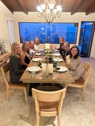 Group of eight women enjoying a celebratory coastal dinner at a long wooden table with wine glasses, sashes and tiaras, chandelier overhead and a large window showing the ocean at dusk.