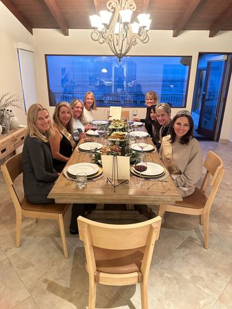Group of eight women enjoying a celebratory coastal dinner at a long wooden table with wine glasses, sashes and tiaras, chandelier overhead and a large window showing the ocean at dusk.