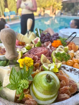 Close-up charcuterie board poolside with sliced pear, fresh figs, cheeses, cured meats, grapes, dried apricots, herbs, edible flowers and a wooden-handled cheese knife at a summer gathering.