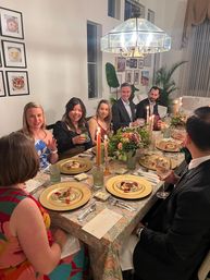 Candlelit dinner party in a cozy dining room — friends gathered around a table with gold chargers, floral centerpiece, chandelier, wine glasses and plated appetizers.