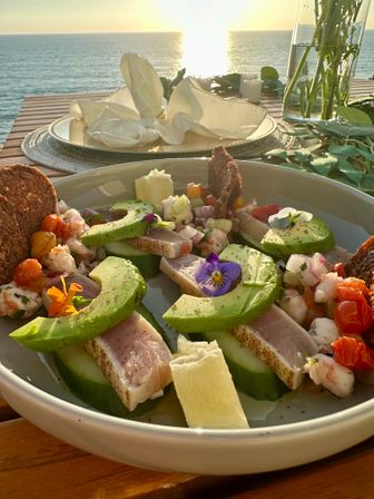 Seared tuna and avocado salad with prawns, cherry tomatoes, cucumber, edible flowers and crisp bread on a wooden table at a seaside sunset — scenic waterfront dining