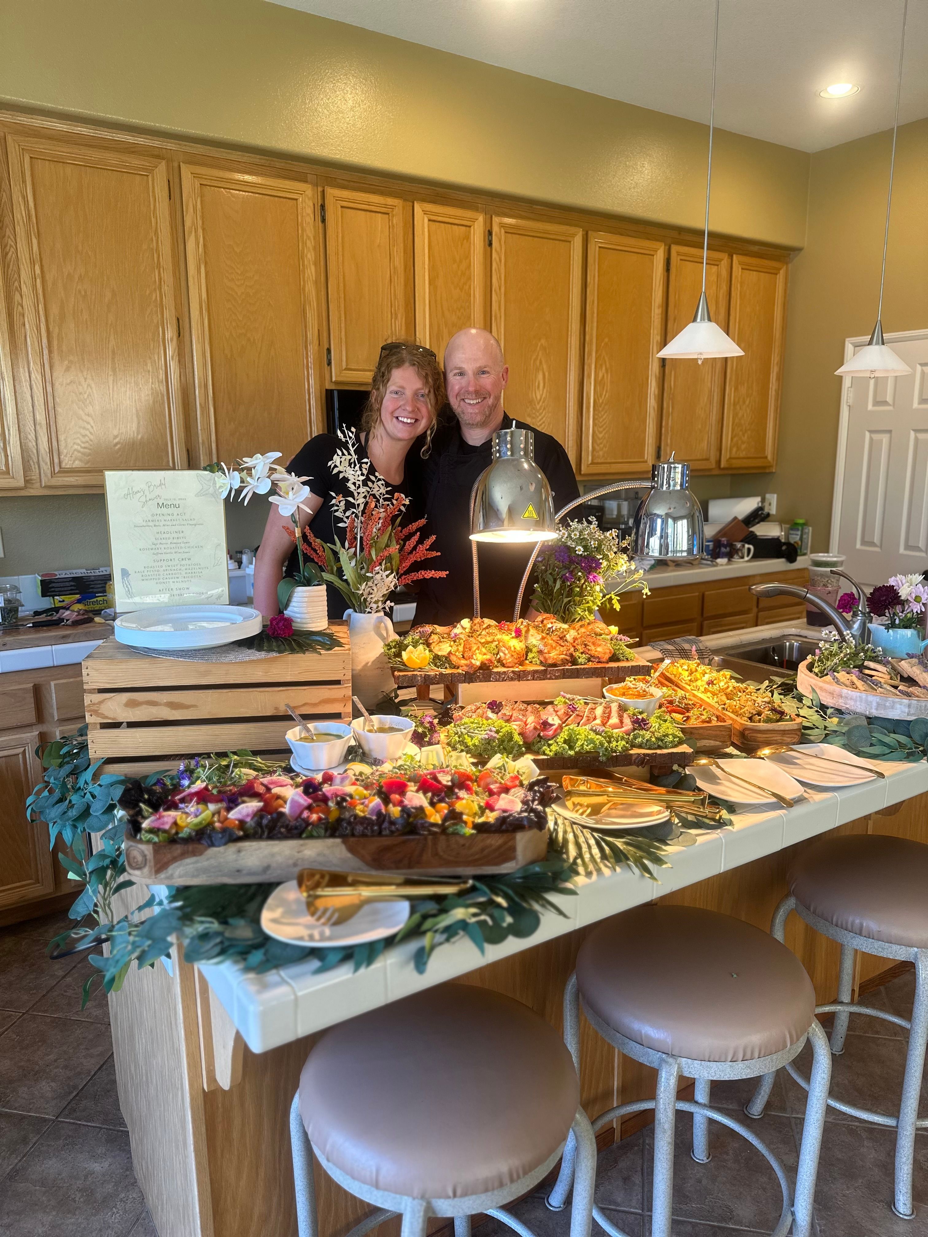 Smiling couple standing behind a vibrant catered spread—charcuterie boards, hot dishes under heat lamps and floral accents—arranged on a home kitchen island with oak cabinets and round counter stools