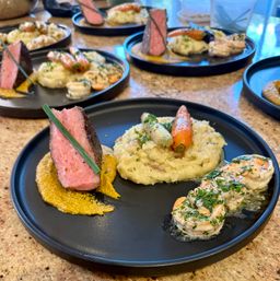 Fine-dining black plate with medium-rare seared steak slice and chive, creamy mashed potatoes topped with glazed baby carrots, herb-butter shrimp, and a golden sauce smear — multiple plated servings on a countertop.