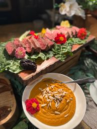 Farm-to-table presentation: sliced medium-rare steak on leafy greens and a rustic wooden board with edible flowers, paired with a bowl of creamy orange dip topped with slivered almonds.